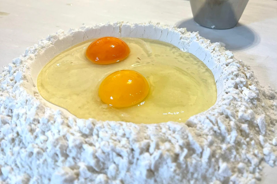 Two raw eggs cracked into a well of flour on a countertop, with a metal cup partially visible in the background.