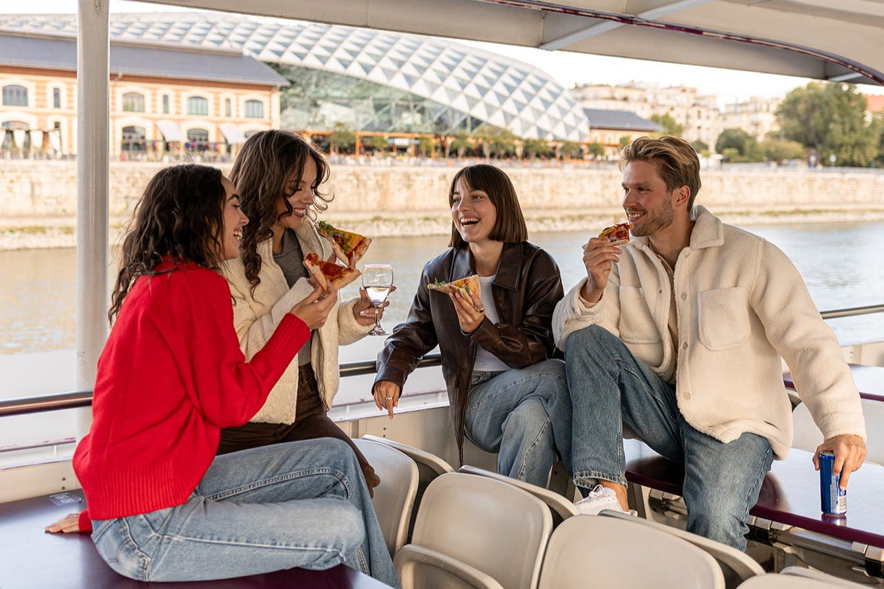 ボートに座ってピザを食べながら会話をする4人。街並みとモダンな建物が背景に。