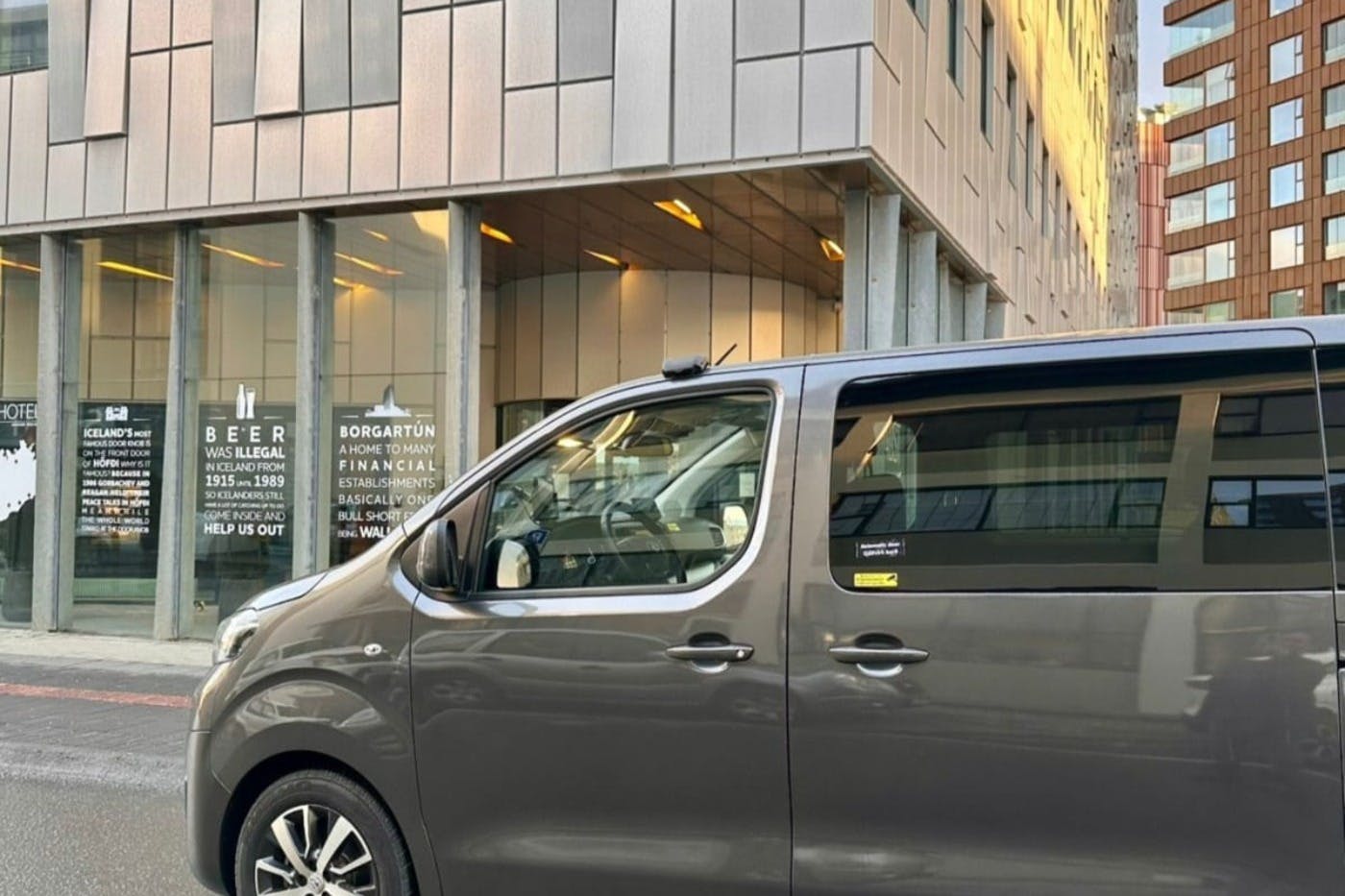 A gray van parked in front of a modern building with large windows and a visible sign listing business details.