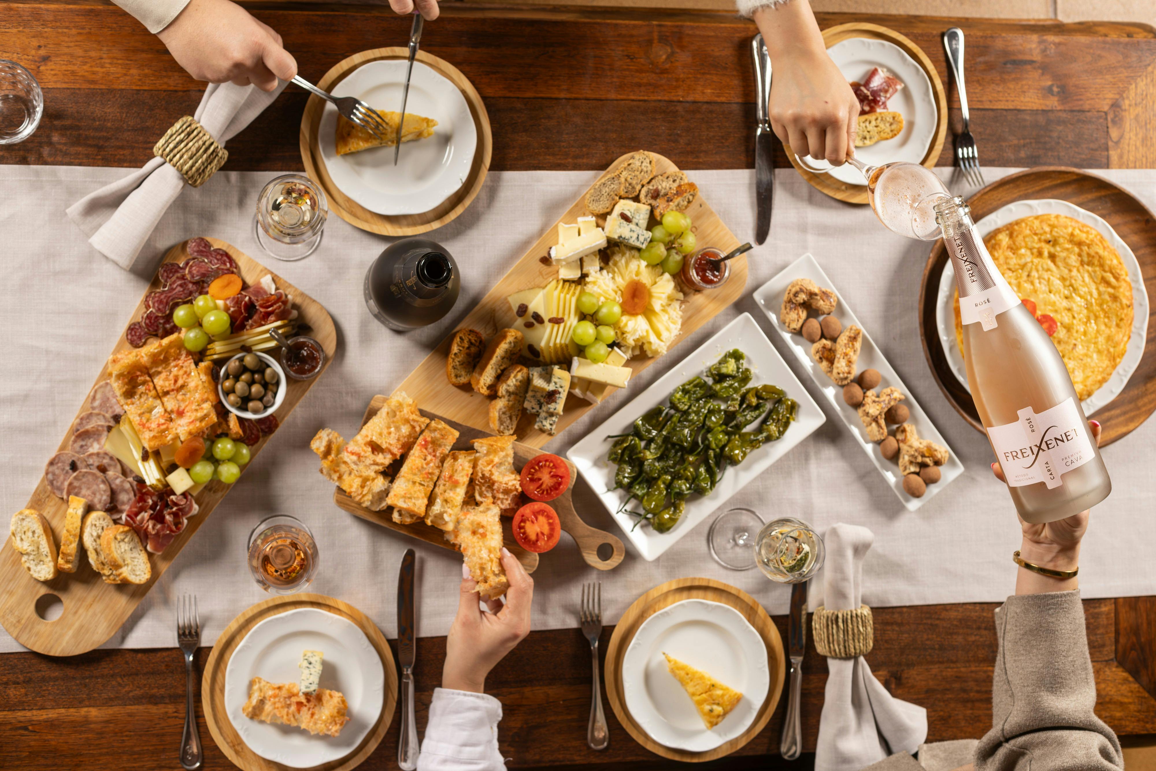 Uma vista aérea de uma mesa de jantar com vários queijos, uvas, pão e outros aperitivos, com quatro pessoas a pegarem na comida.