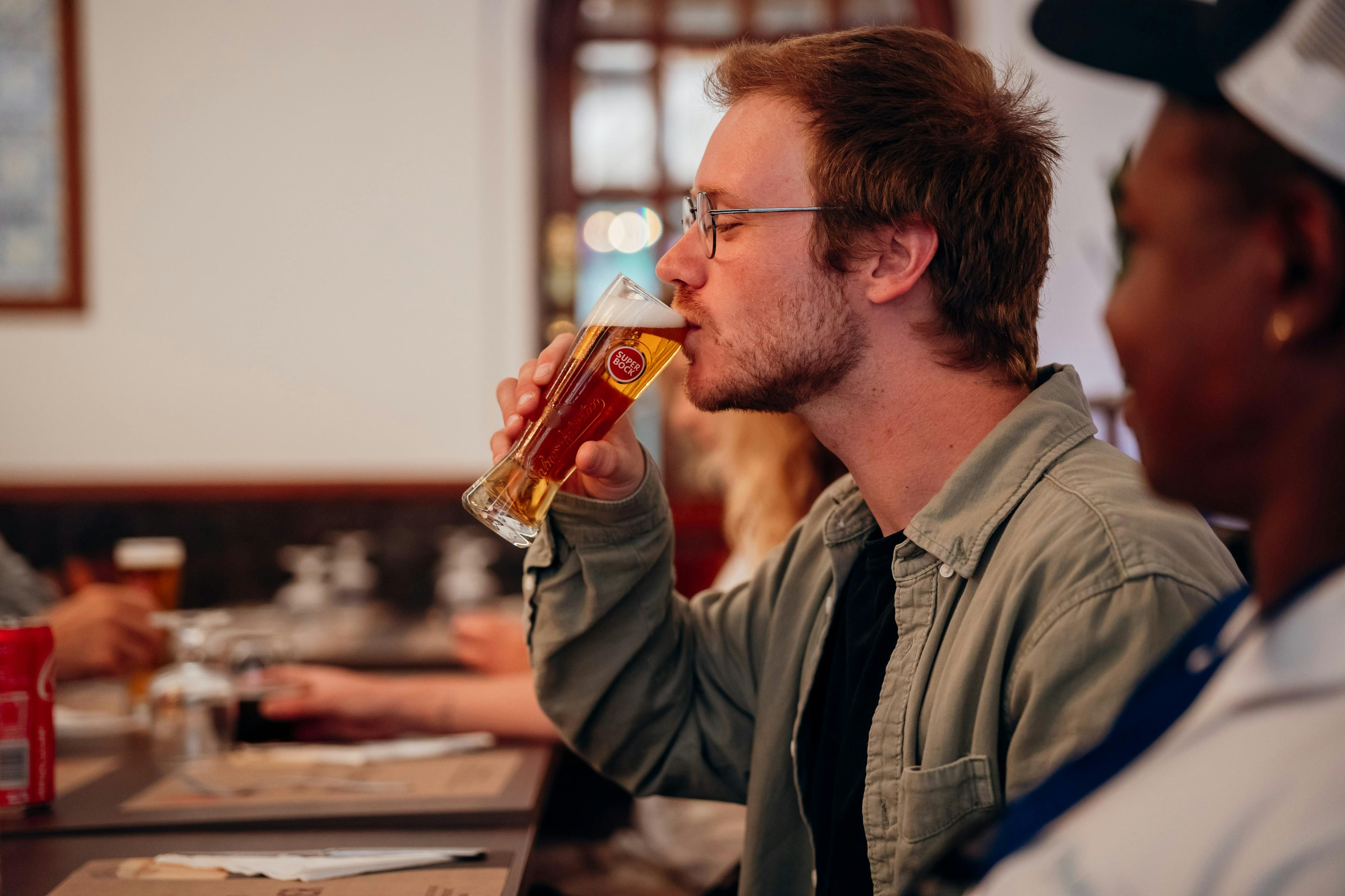 Ein Mann mit Brille trinkt ein Glas Bier in einem ungezwungenen Innenraum mit anderen Personen in der Nähe.