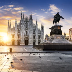 En storslagen katedral vid soluppgången med en ryttarstaty framför, omgiven av historiska byggnader och spridda duvor.