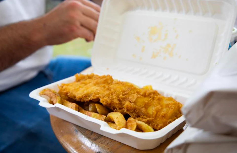 A styrofoam container with fried fish and chips, held by a person in outdoor seating.