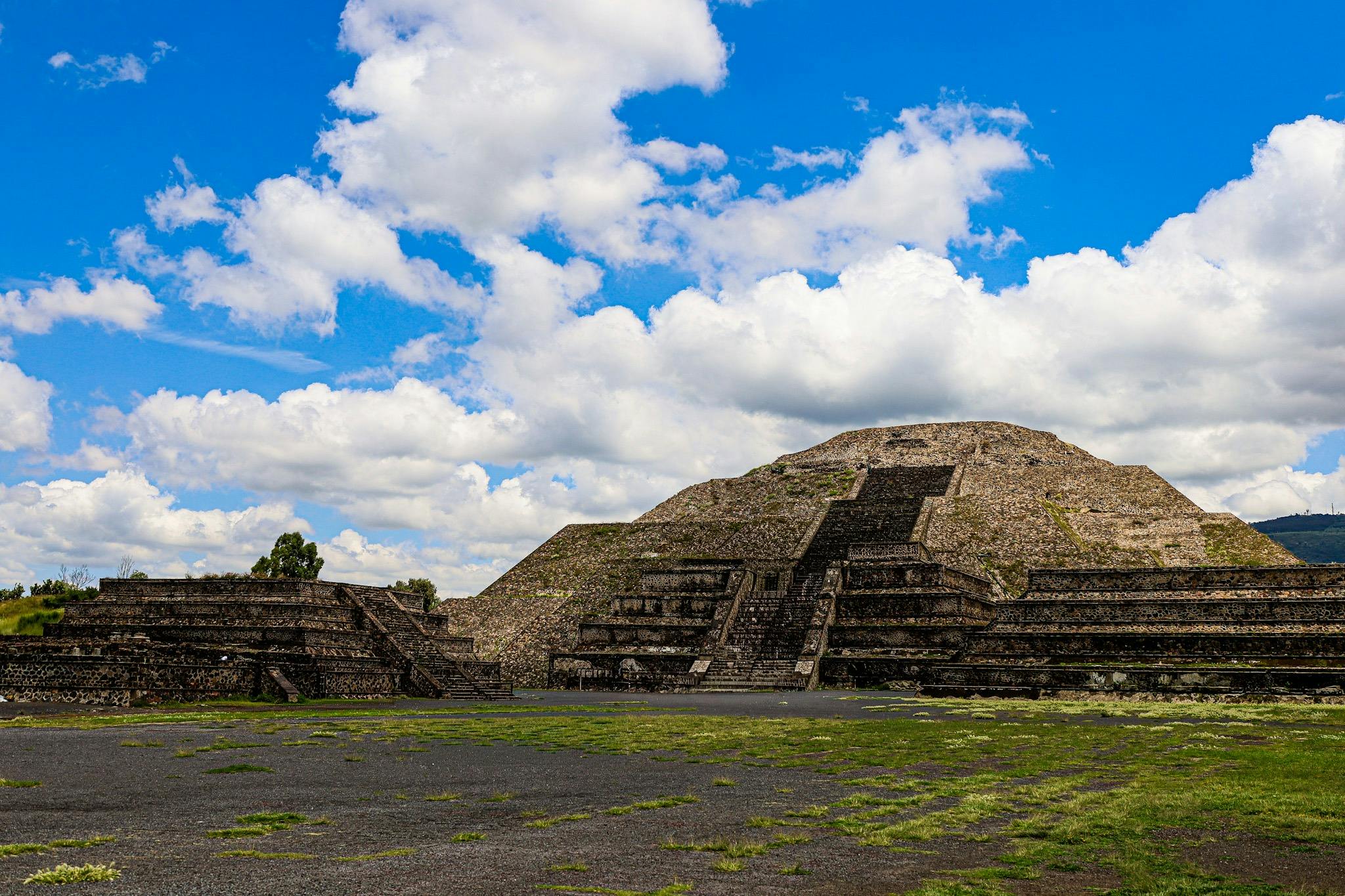A stepped pyramid structure under a bright blue sky with scattered clouds, surrounded by grassy and rocky terrain.