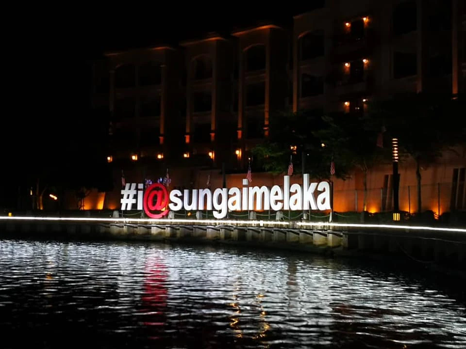 The large illuminated sign "#i@sungaimelaka" is set against the waterfront at night, with a building lit in the background.
