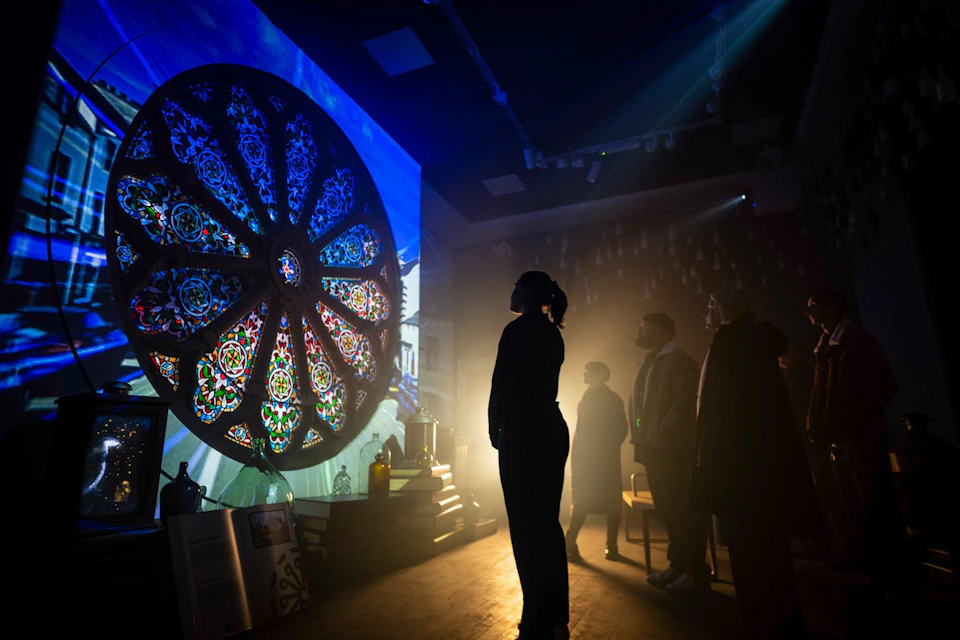 A group of people observe a large, illuminated stained glass projection in a dimly lit room filled with atmospheric lighting.