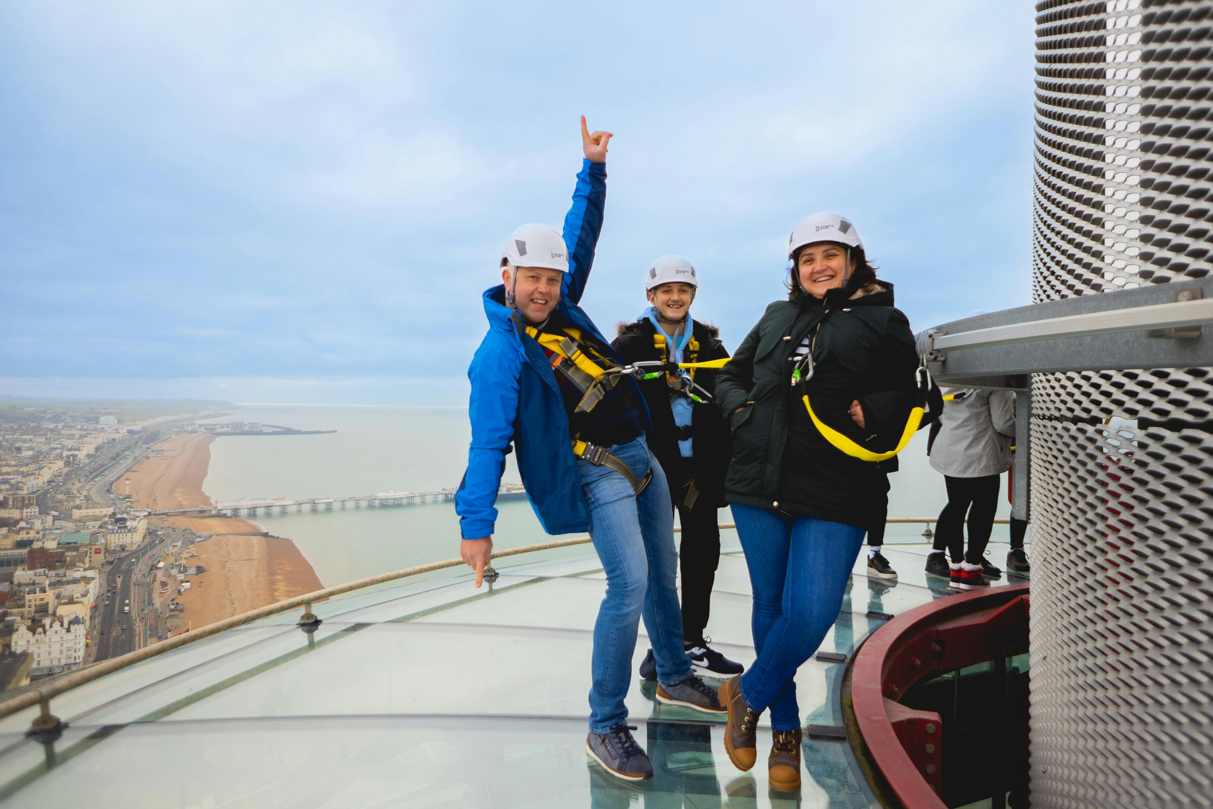 安全ハーネスとヘルメットを着用した3人がガラス張りの展望デッキに立っており、海岸の景色を背景にしています。
