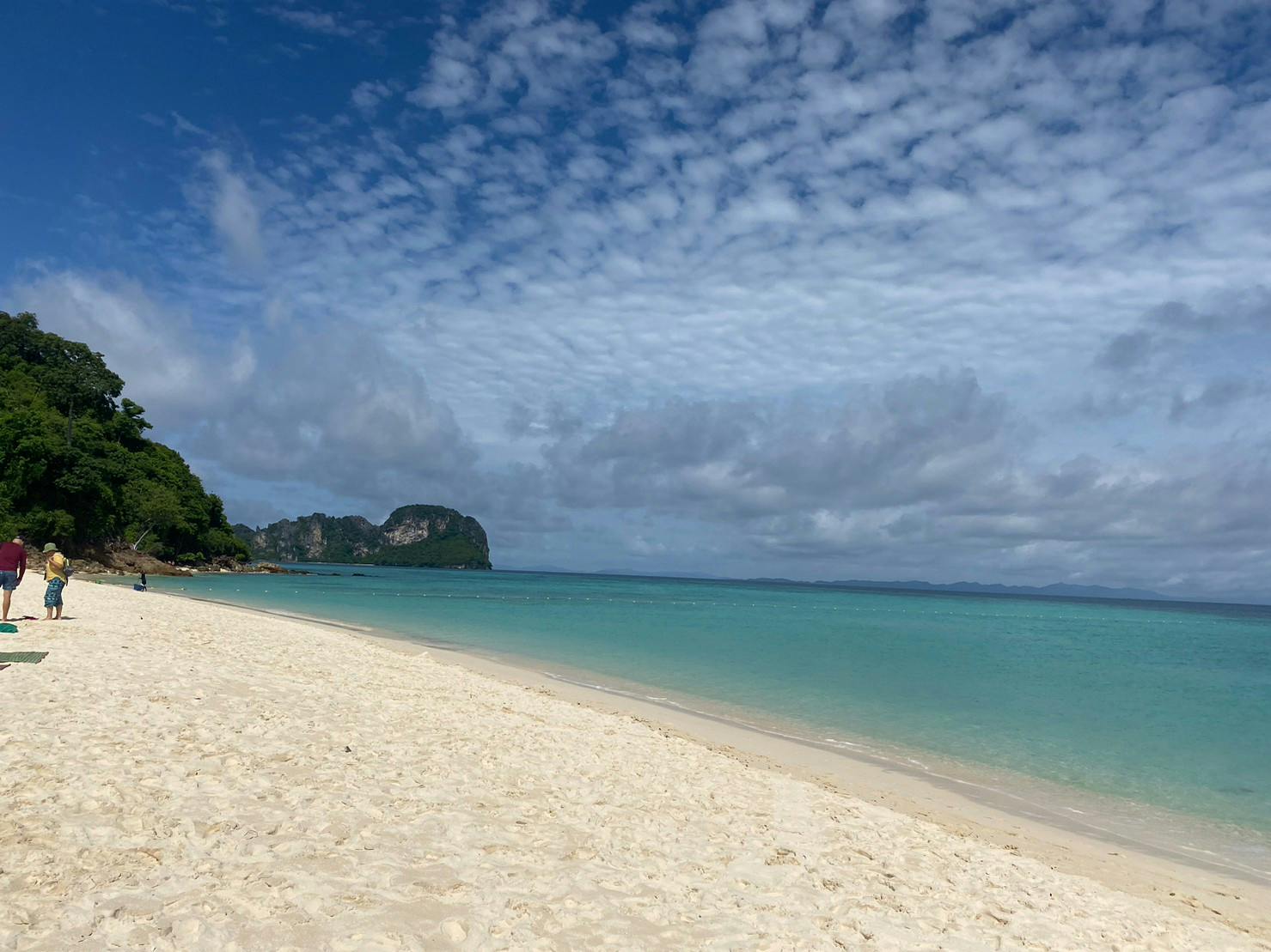Une plage sereine de sable blanc, une eau claire et turquoise et une île montagneuse au loin sous un ciel partiellement nuageux.