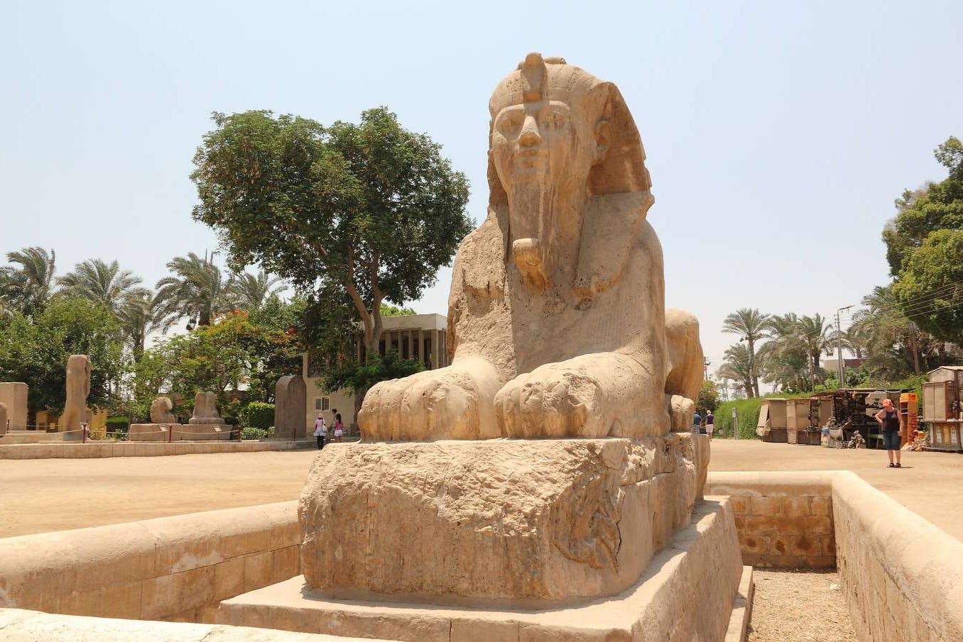 A stone sphinx statue in an outdoor setting with trees and palm trees in the background under a clear sky.