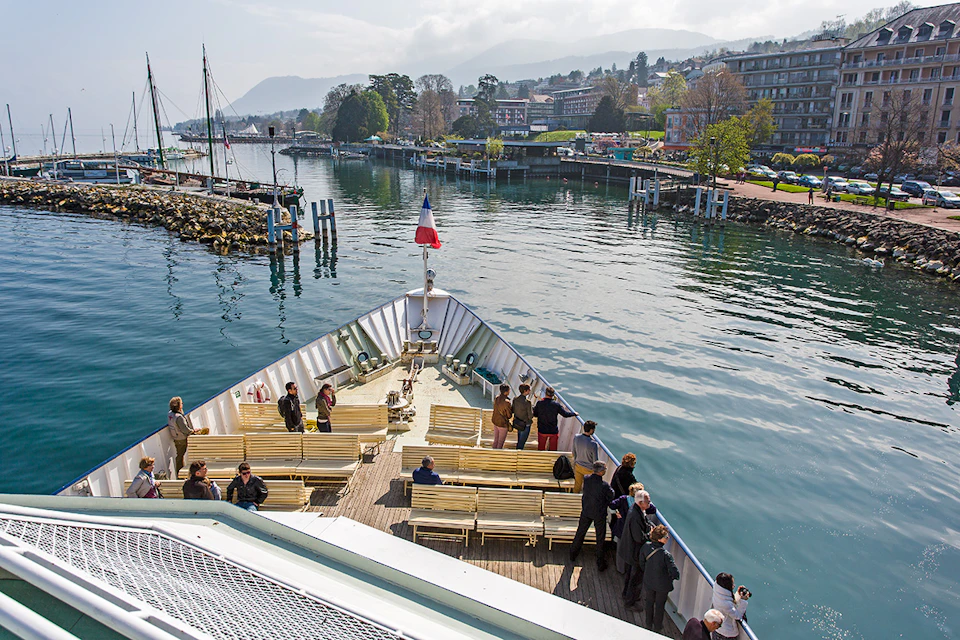 People standing and sitting on the deck of a boat near a shoreline with buildings, trees, and mountains in the background.