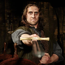 En man med axellångt hår håller ett svärd riktat framåt, framför en träbakgrund och skeppets ratt.