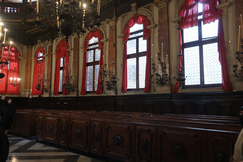 A grand room with large windows, red drapes, wooden pews, and an ornate chandelier hanging from the ceiling.