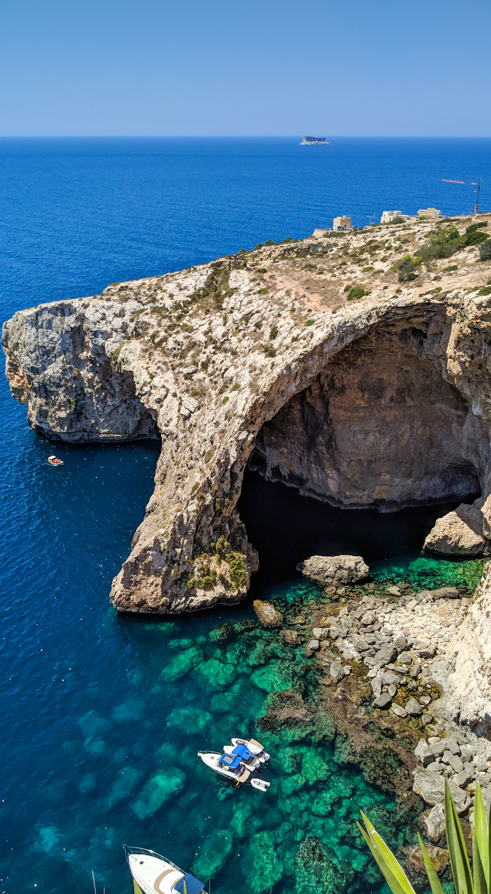 Coastal cliff arch over blue sea with clear water and rocks below; small boat in the sea; distant buildings on the cliff.
