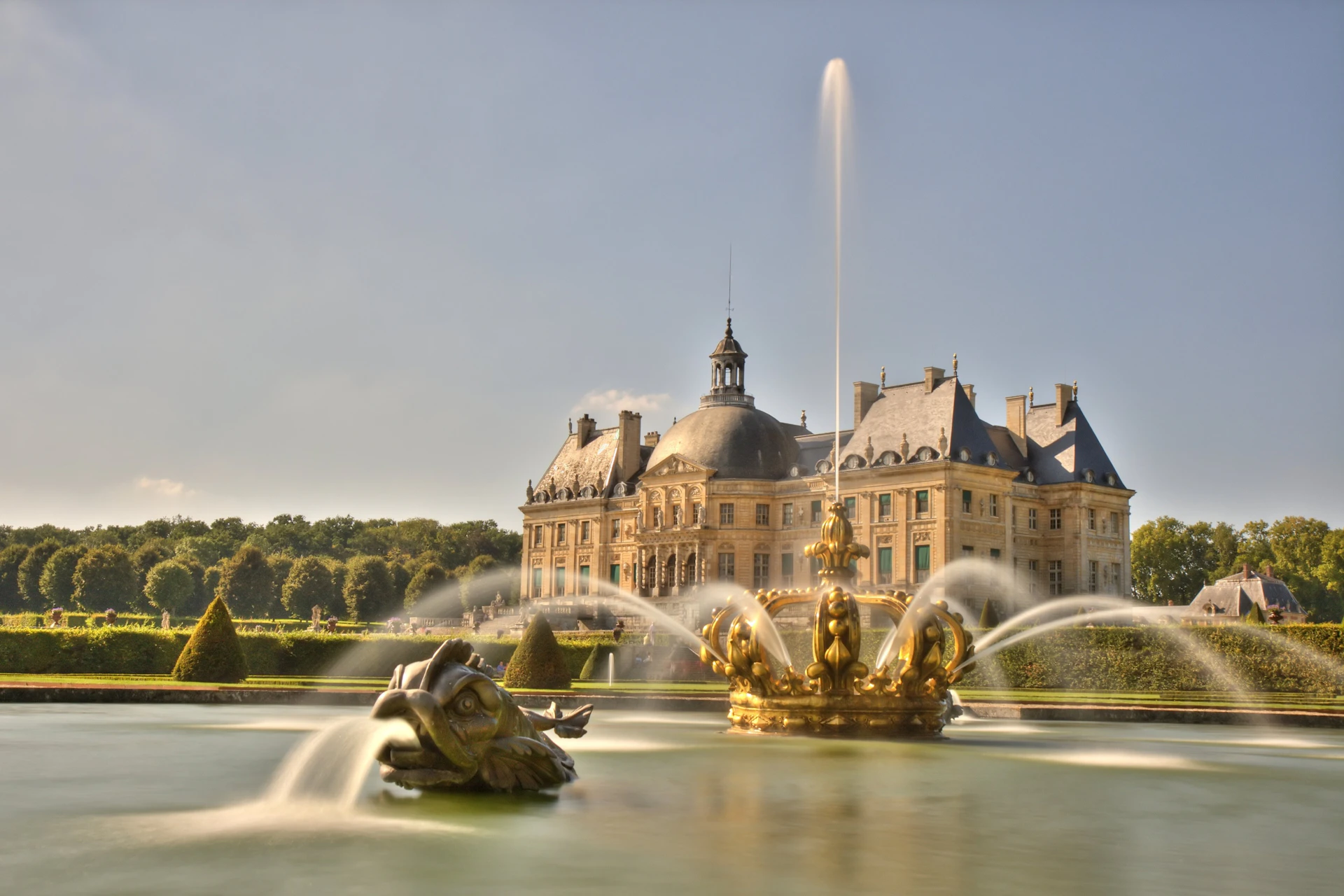 A baroque-style chateau with a fountain in the foreground featuring golden sculptures and water jets.
