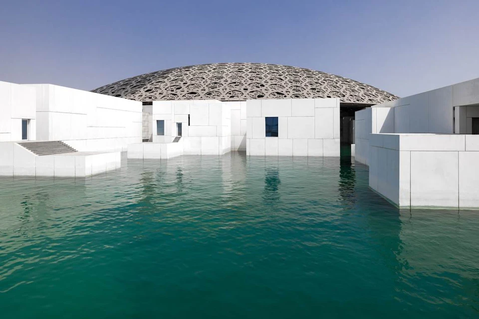 Modern white building with large, intricate dome roof, surrounded by turquoise water under a clear blue sky.