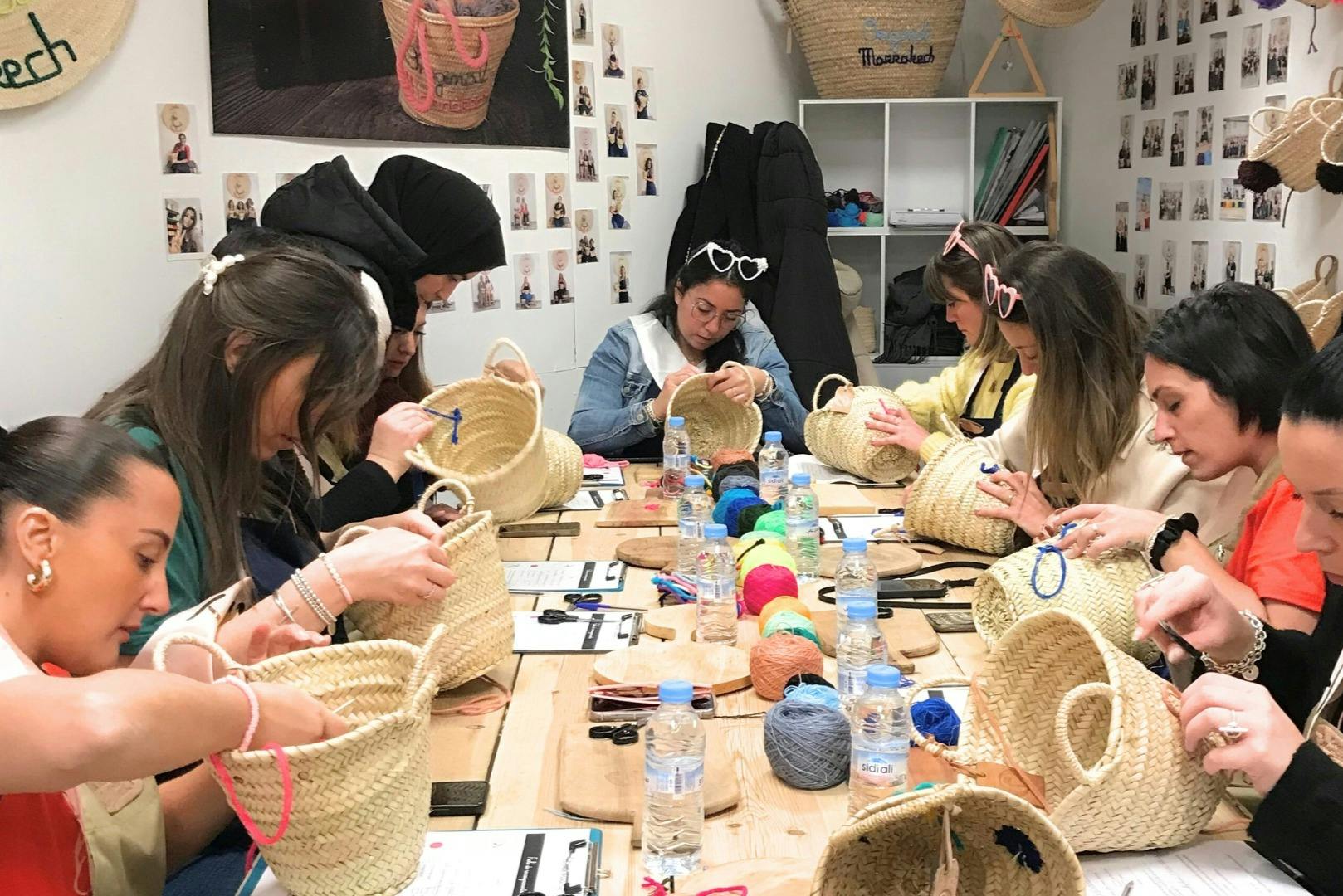 Μια ομάδα ανθρώπων κάθεται γύρω από ένα τραπέζι, υφαίνοντας καλάθια από άχυρο, με εργαλεία και νήματα διάσπαρτα γύρω.