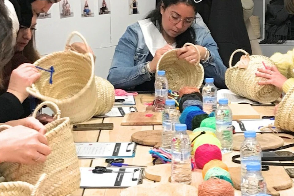 A group of people sitting around a table, weaving baskets from straw, with tools and yarn scattered around.