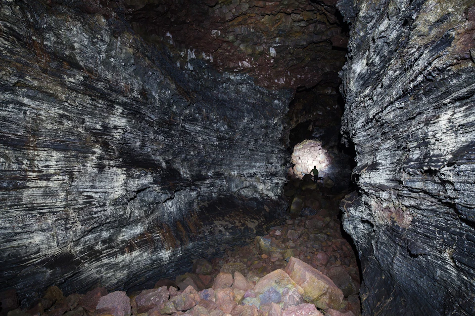 A dark cave with rocky walls and floor, showing a lone person backlit at the entrance.