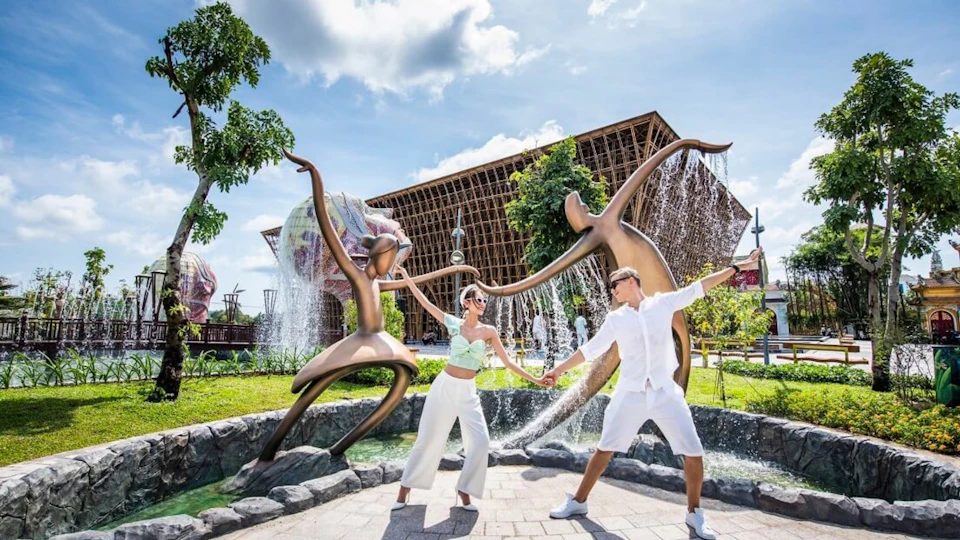 Two people pose in front of a sculpture of dancing figures with water features, set in a sunny outdoor environment.