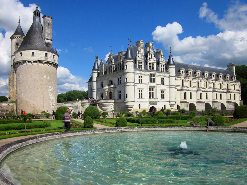 A grand white castle with turrets and detailed architecture, surrounded by gardens and a fountain under a blue sky.