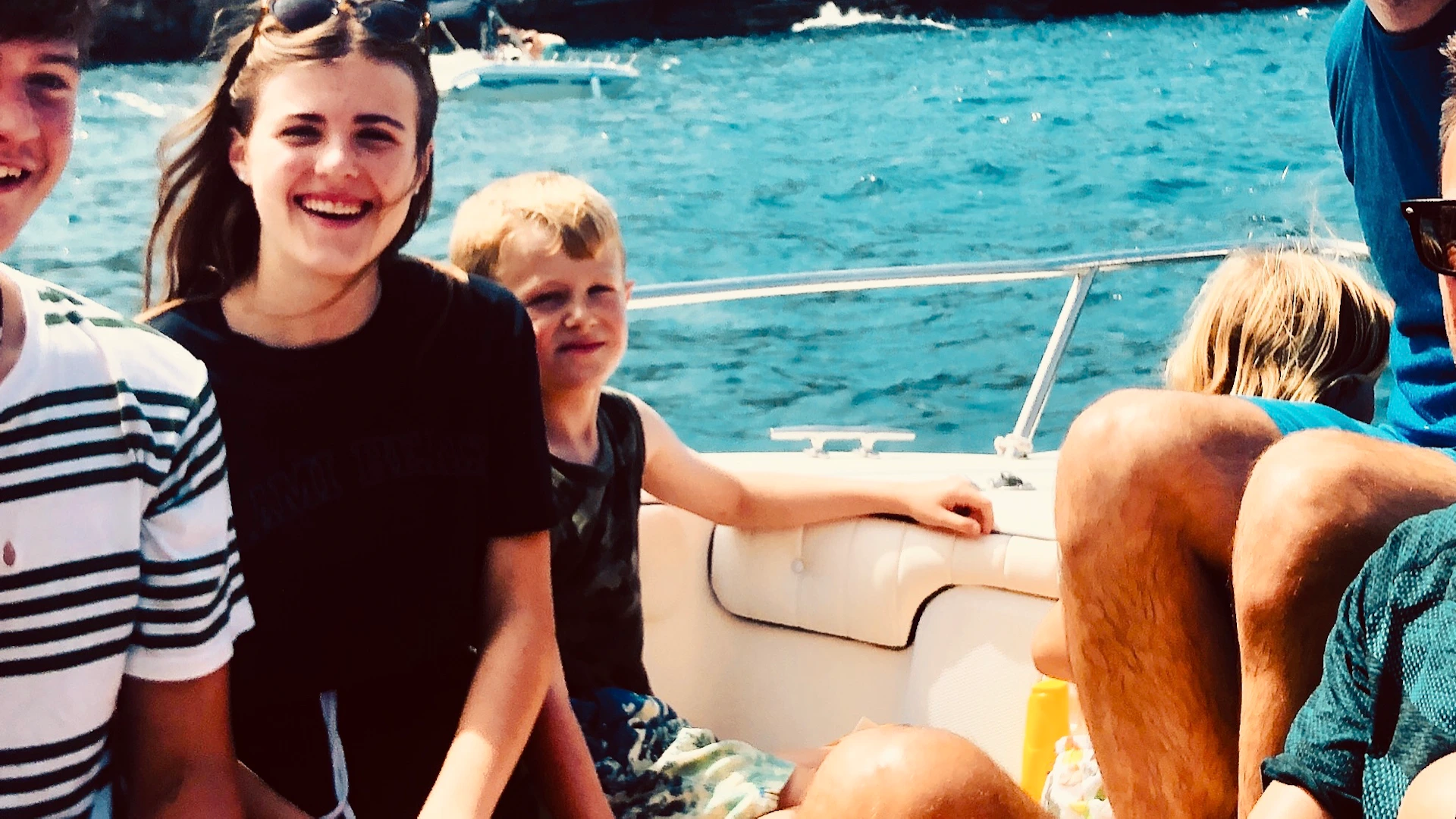 A group of people smiling and sitting on a boat in front of a rocky coastline with buildings, under a partly cloudy sky.