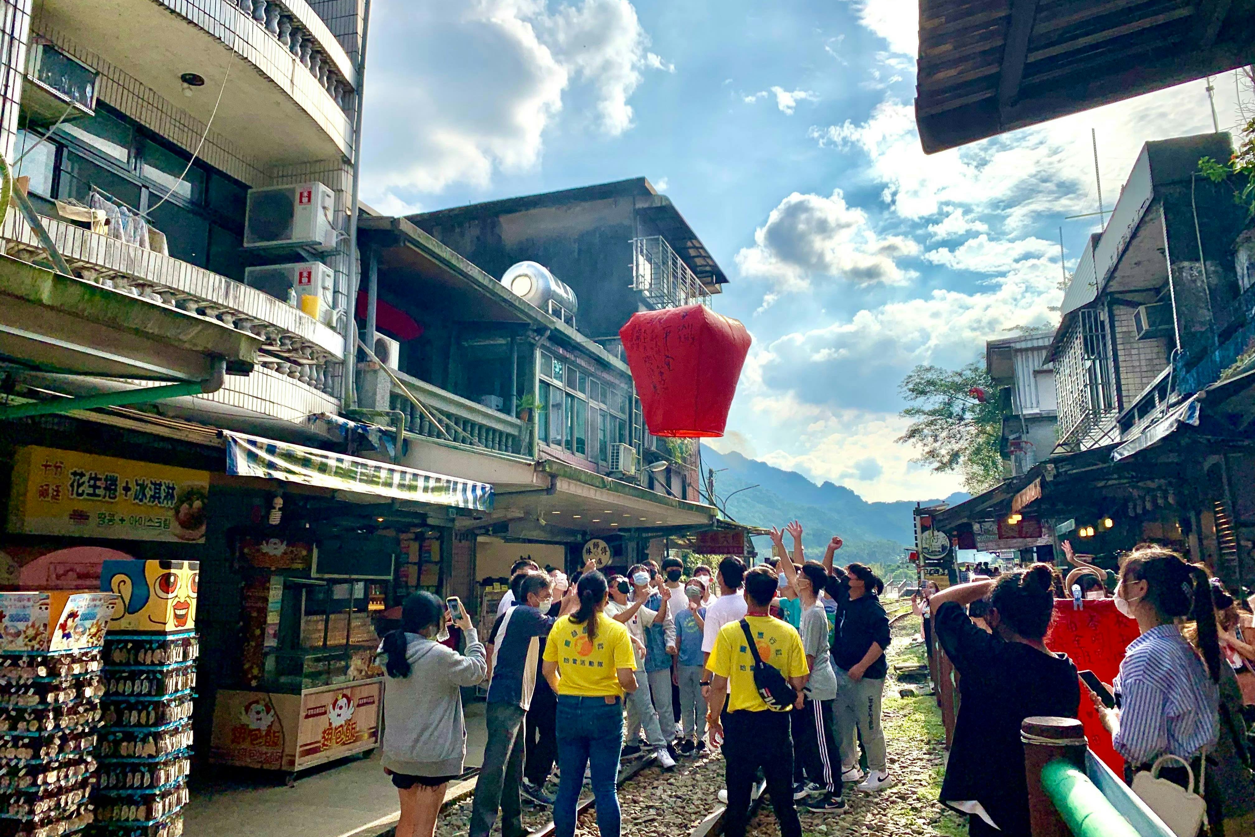 A group of people release a red lantern into the sky in a narrow street lined with buildings under a partly cloudy sky.