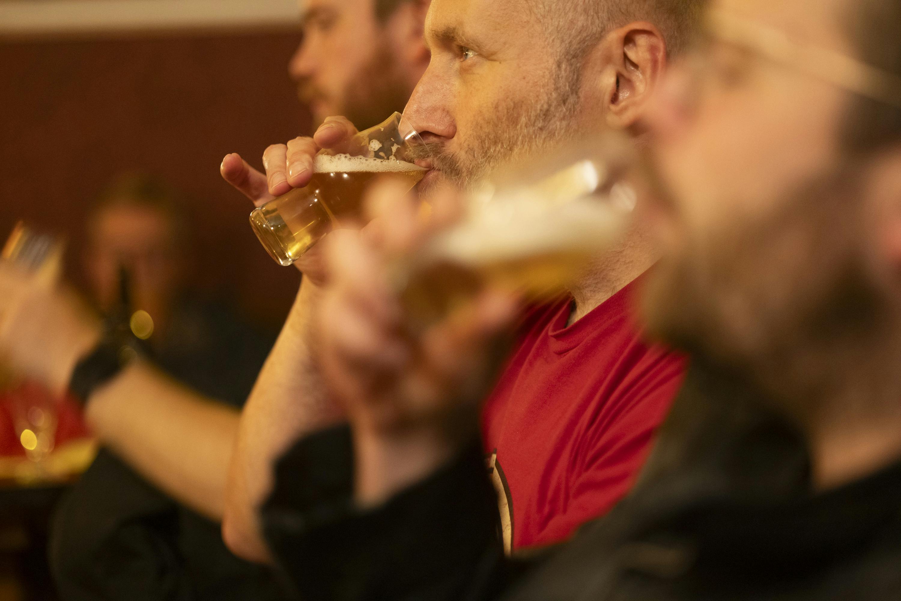 Folk drikker øl i svagt oplyste omgivelser; fokuser på en person i en rød skjorte, der nipper fra et glas.