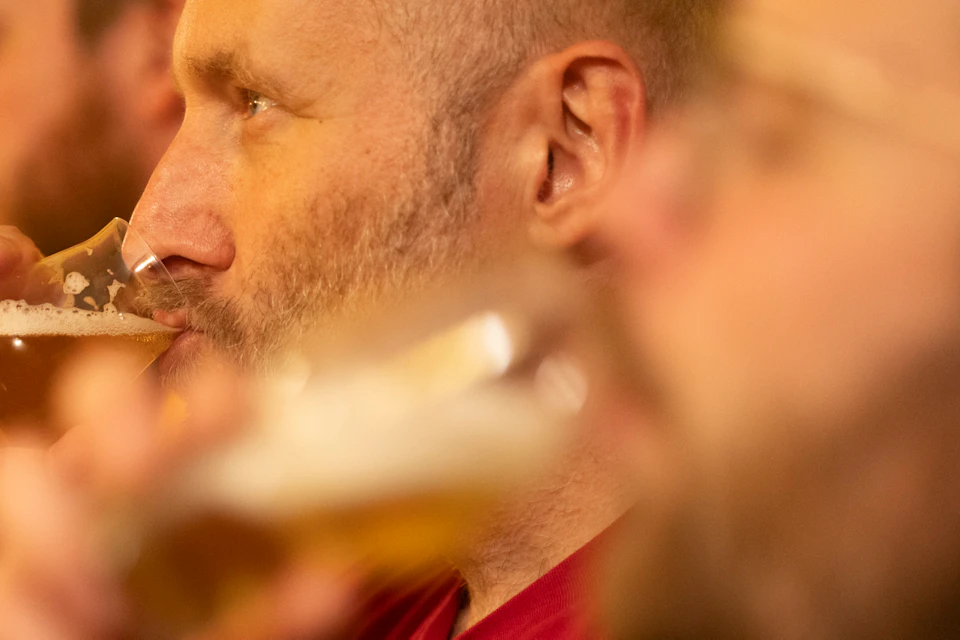 People drinking beer in a dimly lit setting; focus on a person in a red shirt sipping from a glass.