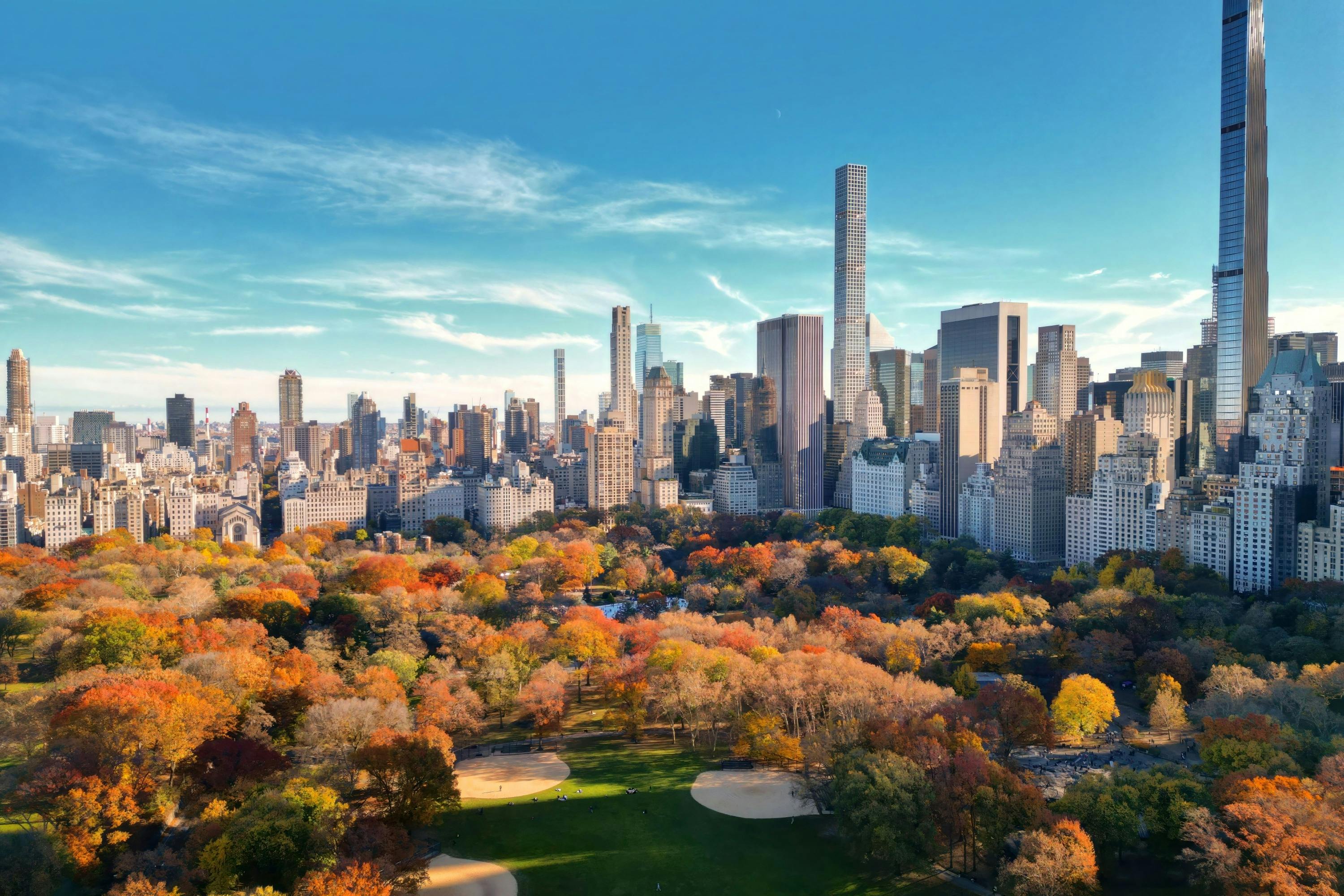 Aerial view of a park with vibrant autumn foliage, two sand baseball fields, and a city skyline under a clear blue sky in the background.