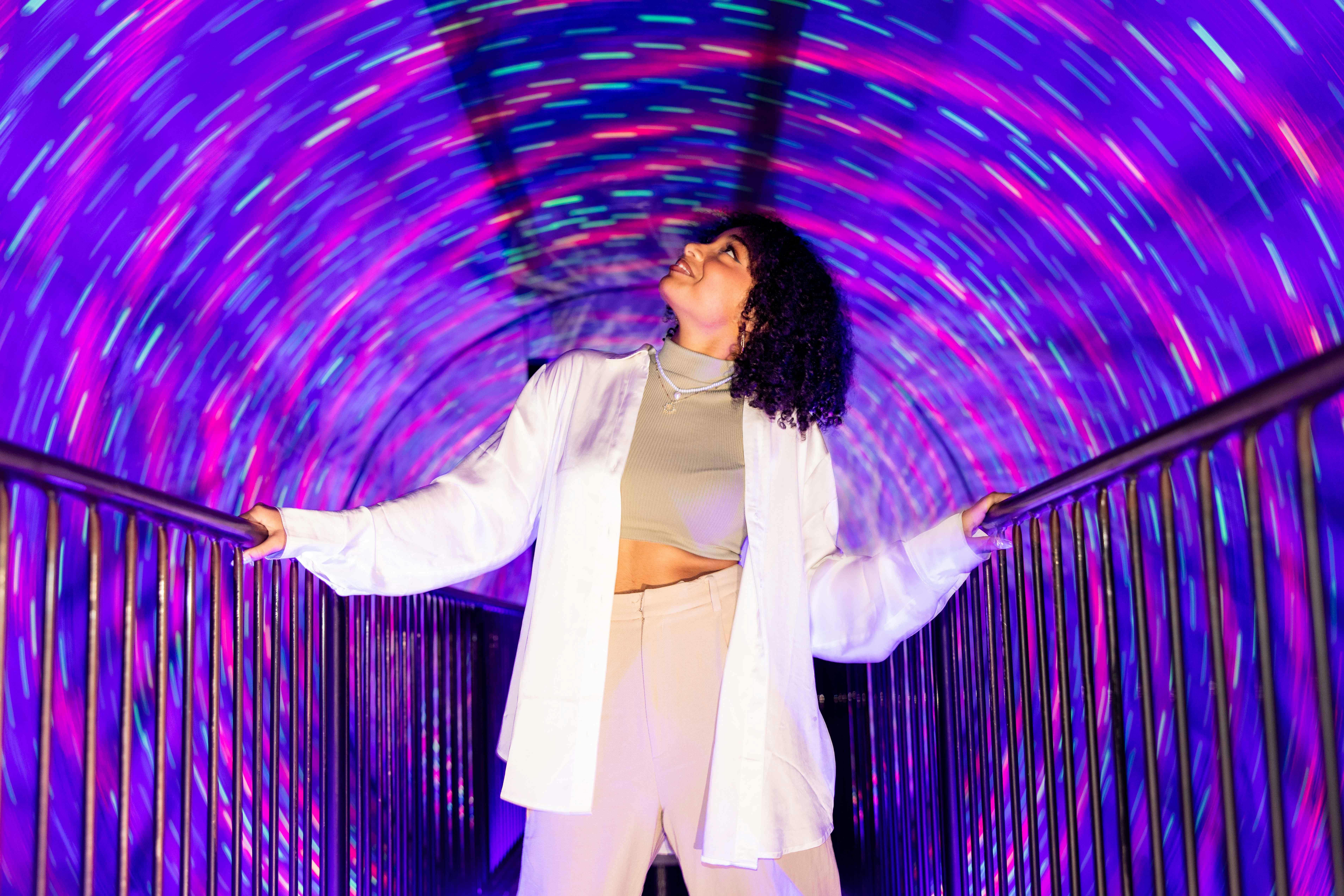 A person stands on a bridge inside a tunnel with swirling purple and blue lights, looking upward with a smile.