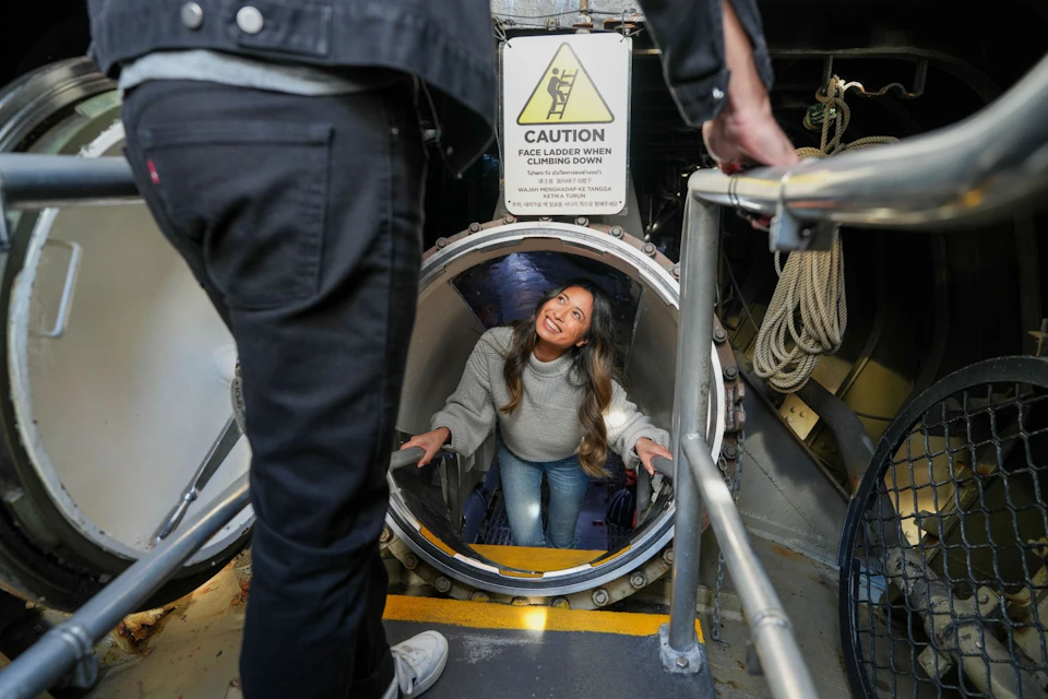 A woman smiling as she climbs up through a circular hatch in a mechanical setting, with caution signage visible above.