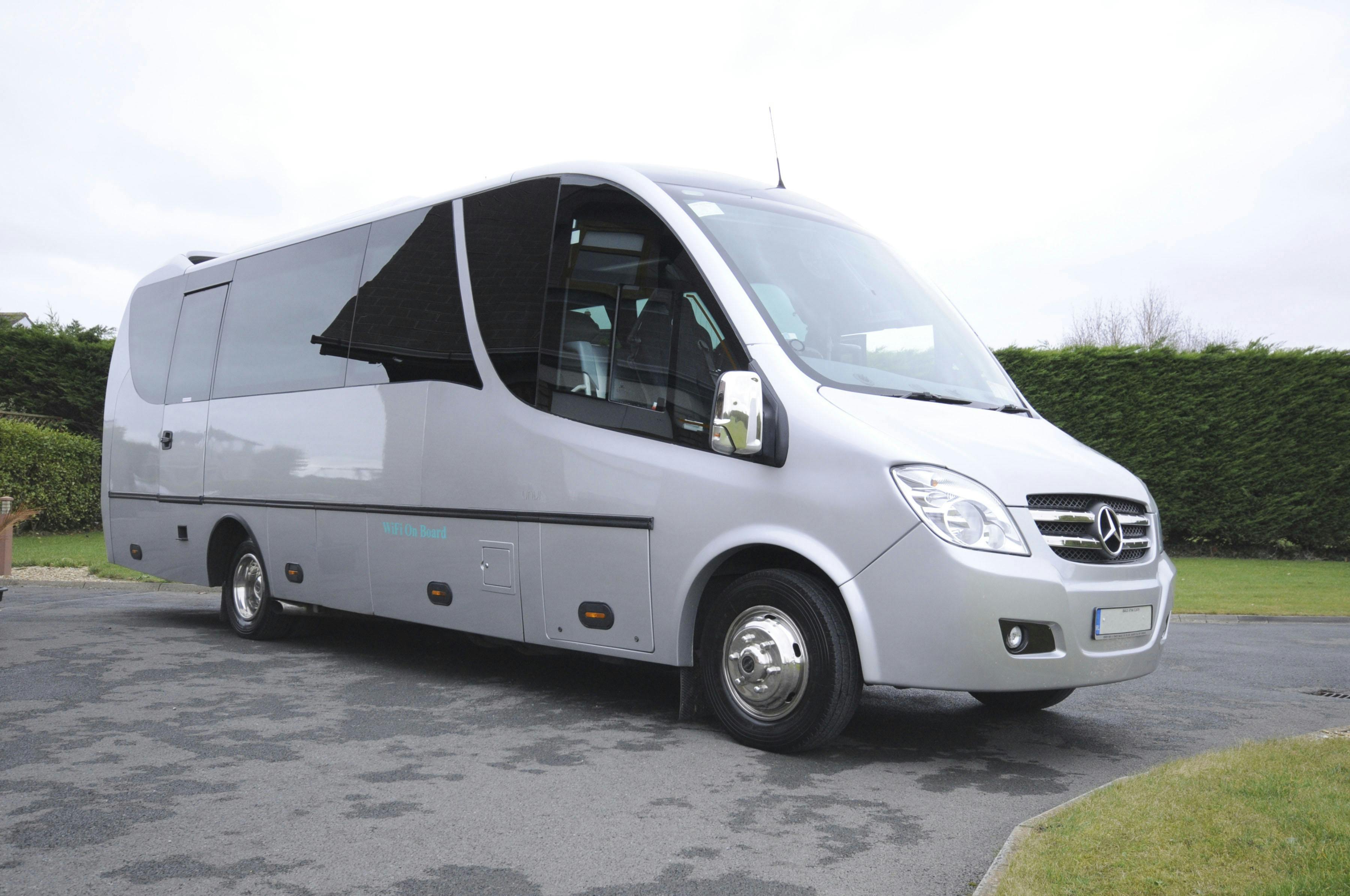 一辆银色梅赛德斯-奔驰小巴停在阴天下的铺面区域，背景是绿色植物。