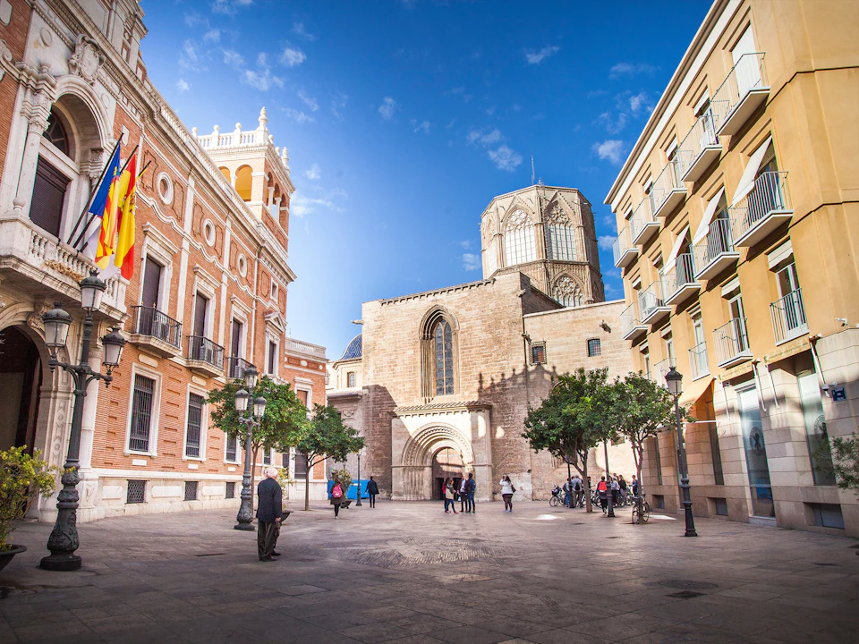 People walking through a plaza surrounded by historic buildings with flags, trees, and a church in the background under a blue sky.