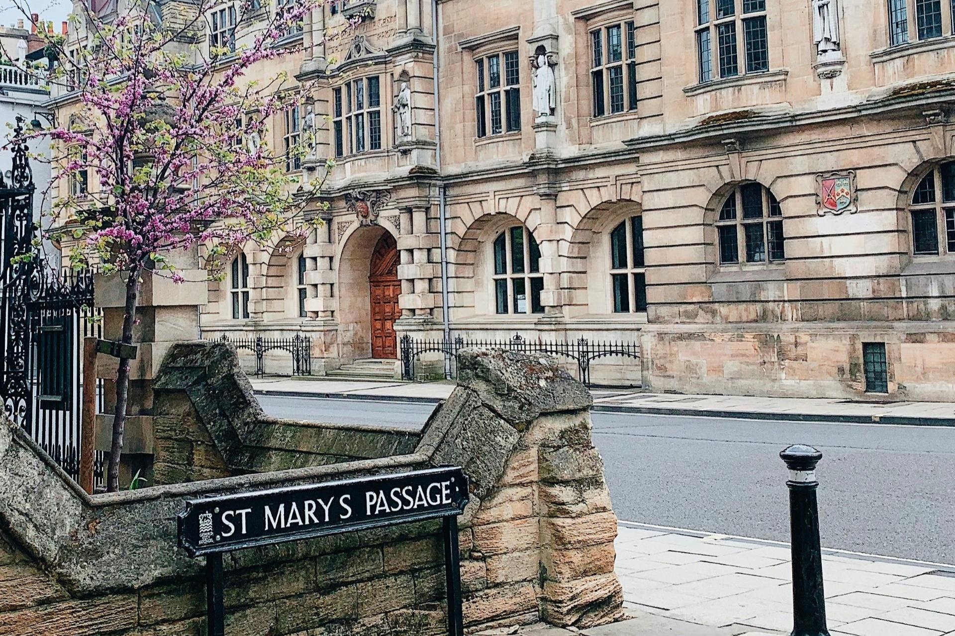 A street view of a historic stone building with arched windows and a sign for St Mary's Passage in the foreground.