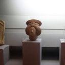 Small stone sculpture of a head with a headdress, displayed on a pedestal, under focused lighting in a museum setting.