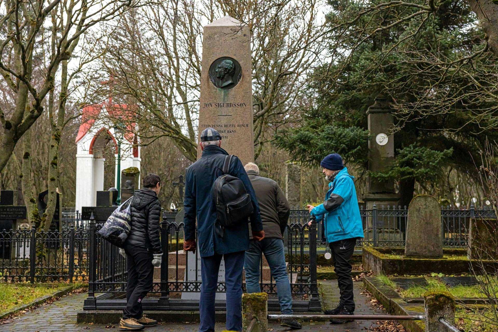 Four people stand around a tall, brown monument in a cemetery, reading the inscriptions. Trees and headstones are visible in the background.