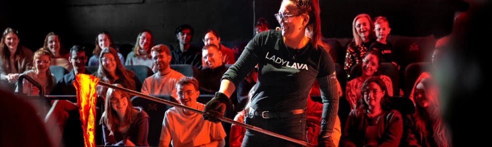 薄暗い劇場に座っている観客が、棒に溶けたガラスをかけた吹きガラスを見せている女性。