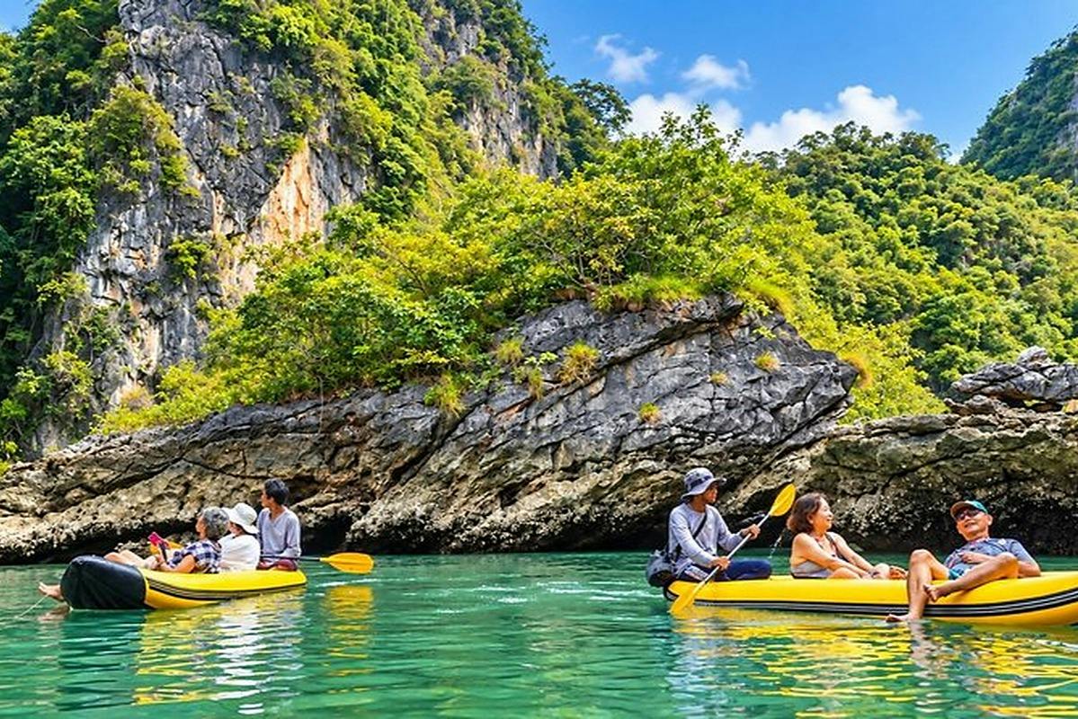 People kayaking on calm green water near rocky cliffs covered with lush vegetation under a partly cloudy sky.