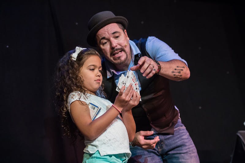 A man in a bowler hat guides a young girl holding playing cards during a magic trick. Both are focused on the cards.