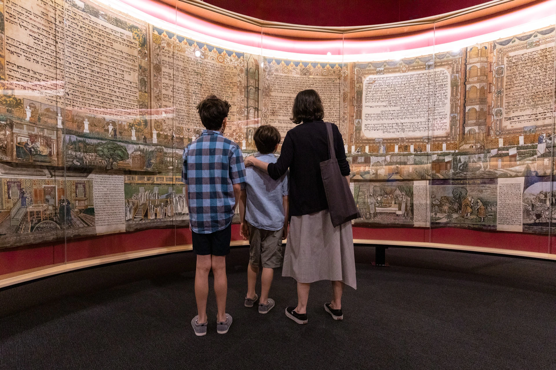 Three people stand in front of a large, illuminated manuscript display in a museum setting.
