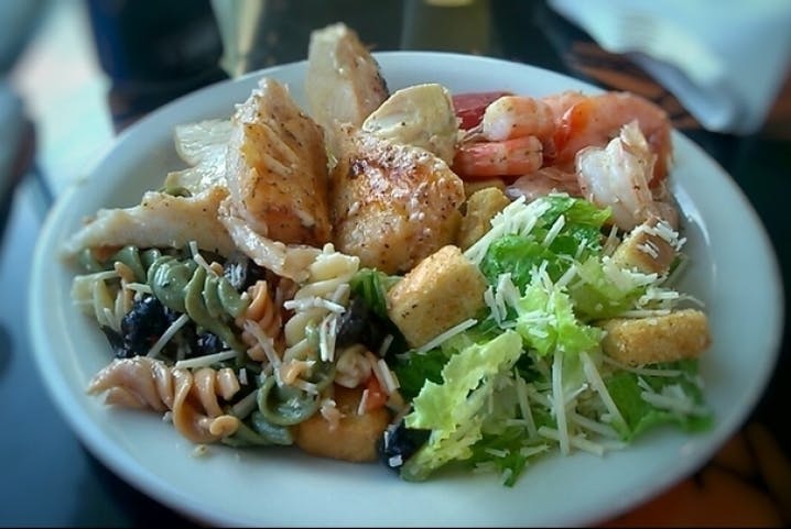 A plate with grilled chicken, shrimp, pasta salad, and Caesar salad with croutons and cheese.