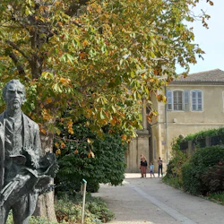 A path lined with greenery leads to an old building. A statue of a man holding a book is in the foreground. Two people stand in the distance.