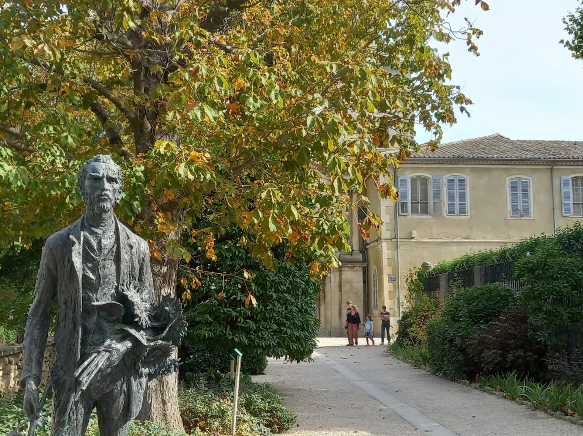 A path lined with greenery leads to an old building. A statue of a man holding a book is in the foreground. Two people stand in the distance.