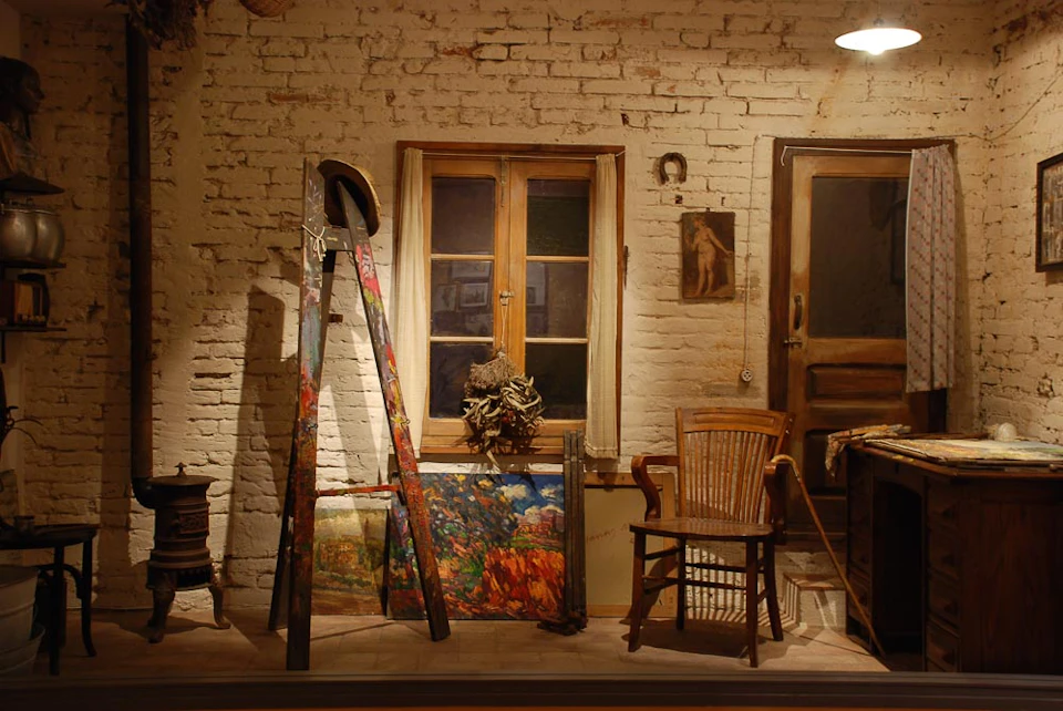 An artist's studio with a paint-splattered easel, framed paintings, a wooden chair, and a window on a brick wall.