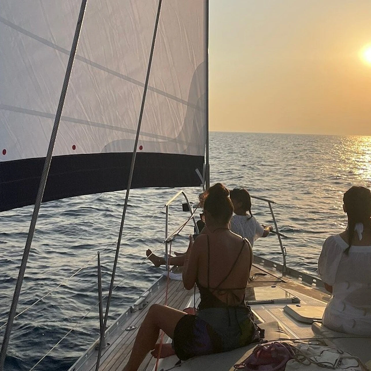 Group of people sitting on the deck of a sailboat at sunset, with the sails raised and the sea extending to the horizon.