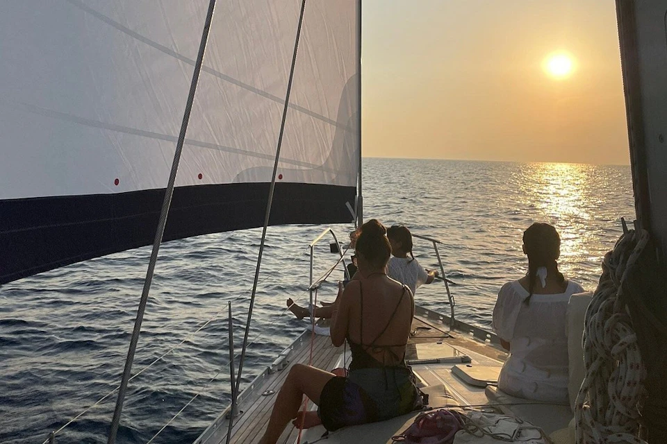 Group of people sitting on the deck of a sailboat at sunset, with the sails raised and the sea extending to the horizon.
