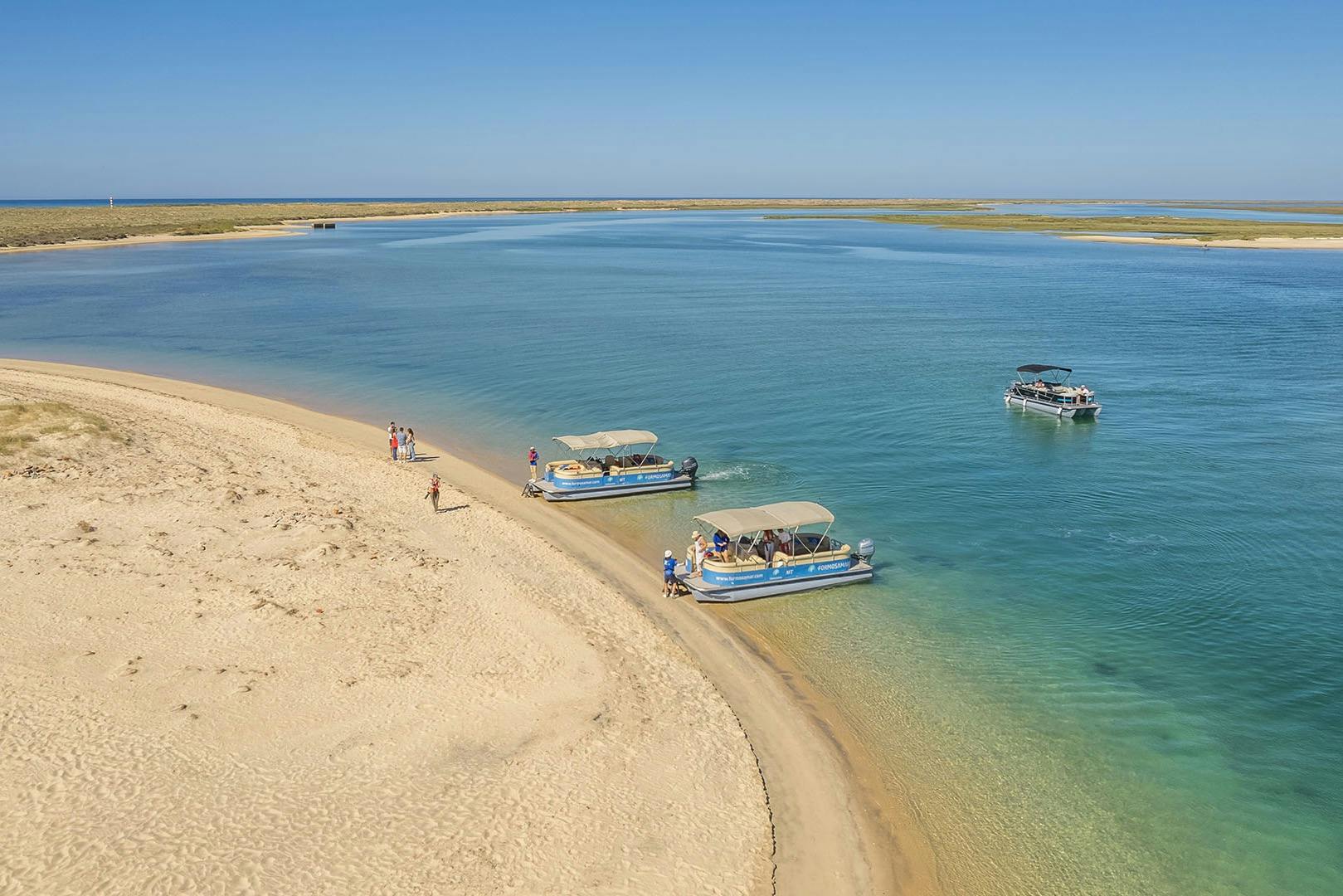 Лодки Formosamar пришвартовались вдоль песчаного берега с людьми, исследующими пляж, и спокойной голубой водой под чистым небом.