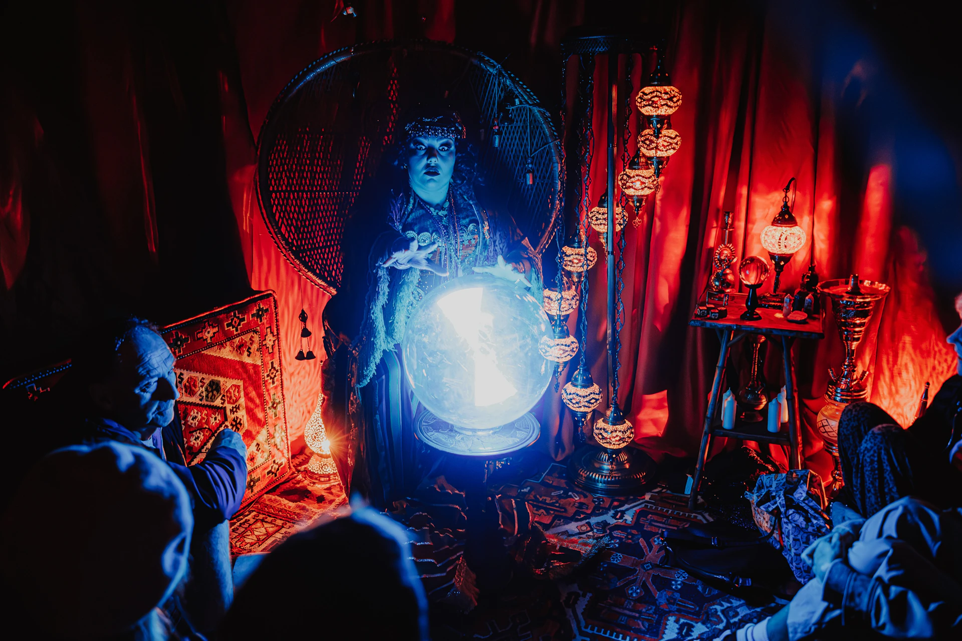 A person in dramatic lighting gazes into a glowing crystal ball in a dimly lit room with ornate decor and hanging lamps.