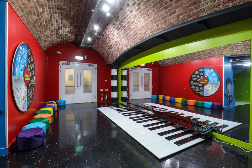 A brightly colored room with a large floor piano, red walls, brick ceiling, and colorful seating along the walls.
