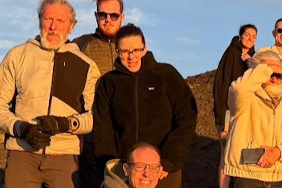 A group of people posing outdoors on a sunny hilltop with a blue sky and scattered clouds in the background.